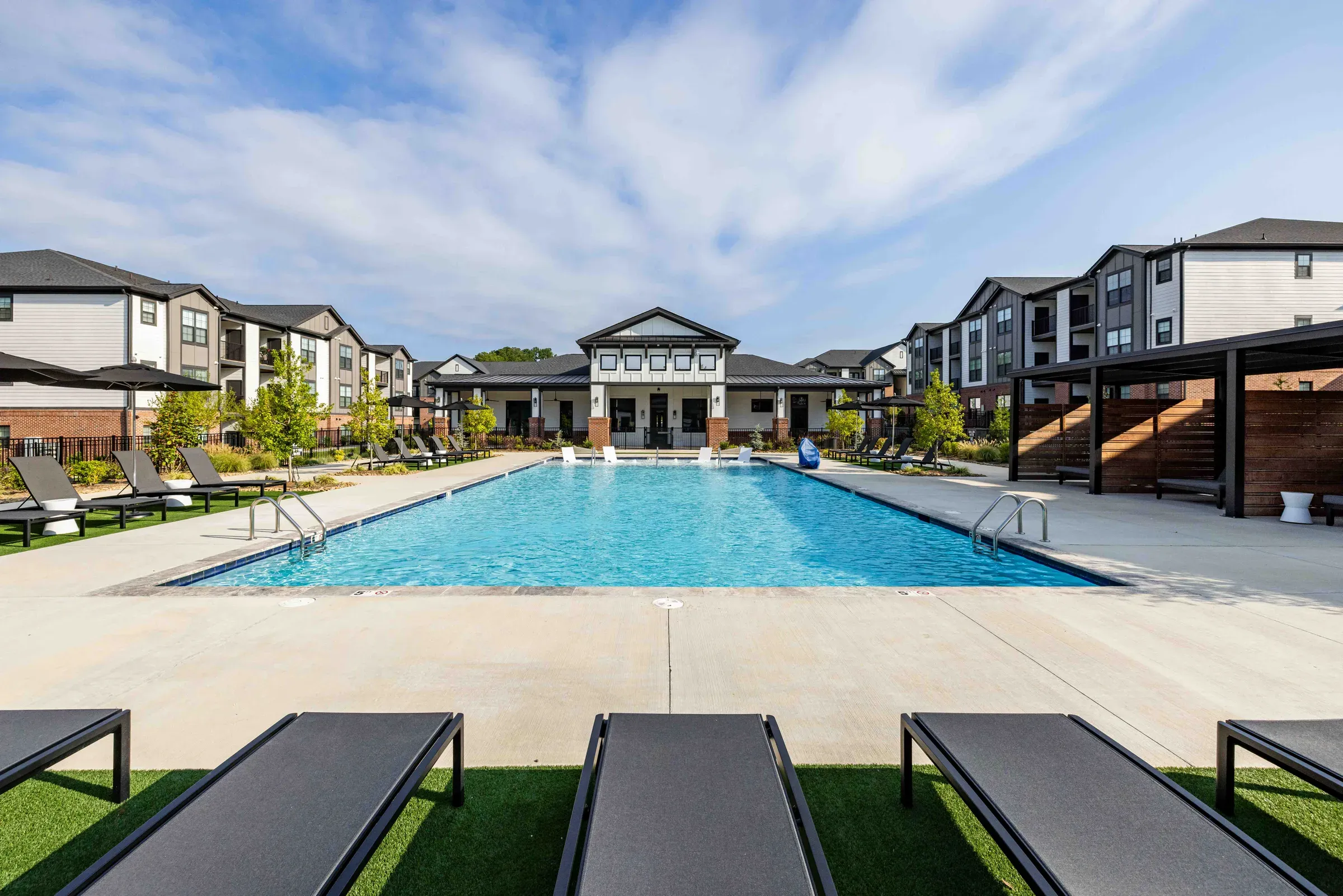 Modern apartment complex with a large outdoor swimming pool, lounge chairs, and surrounding buildings.